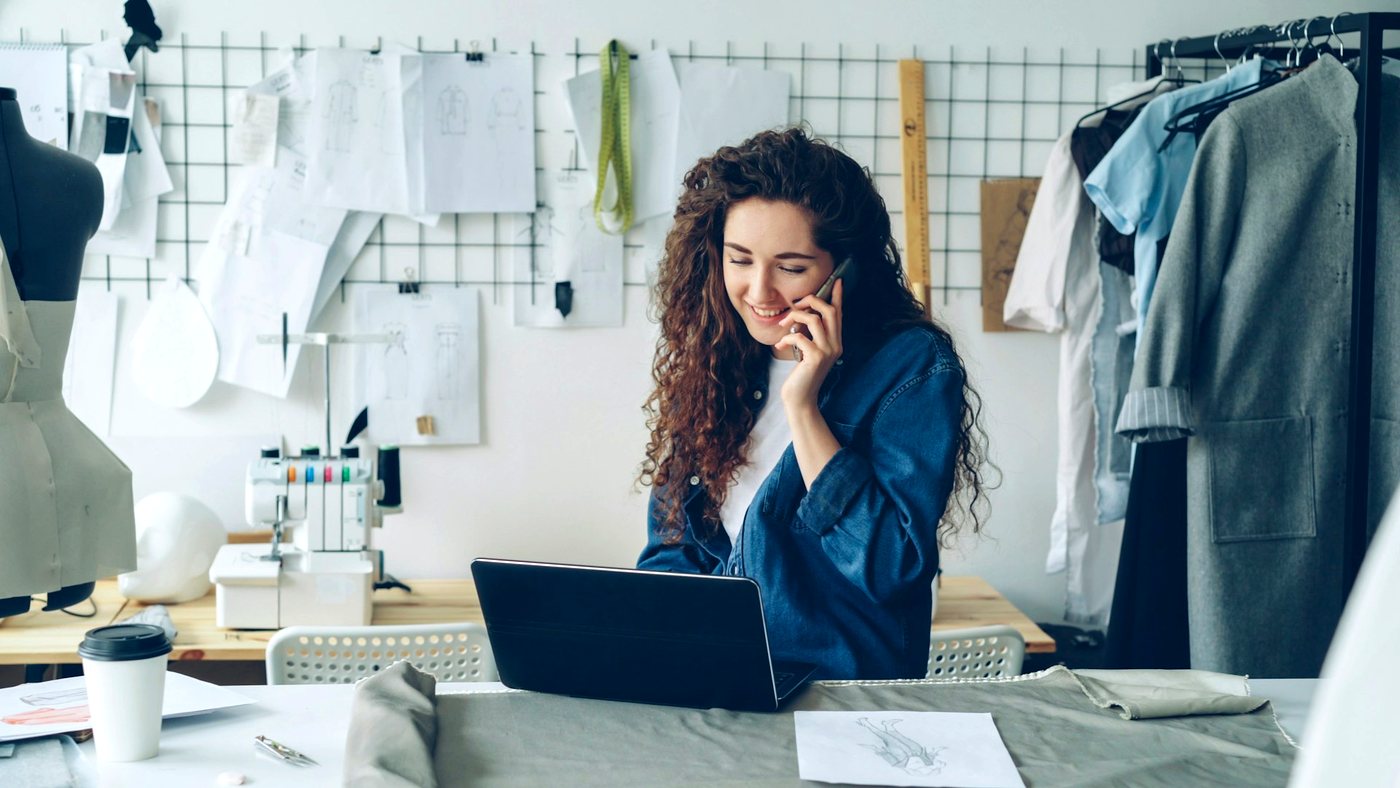 A clothing designer on the phone at her studio worktable, sketches pinned to the wall behind her.